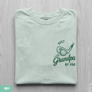 May include: A light green t-shirt featuring a dark green graphic of a golf club and ball with the text "BEST Grandpa BY PAR" printed on the front. The shirt has a crew neckline and a label on the inside collar that reads "COMFORT".