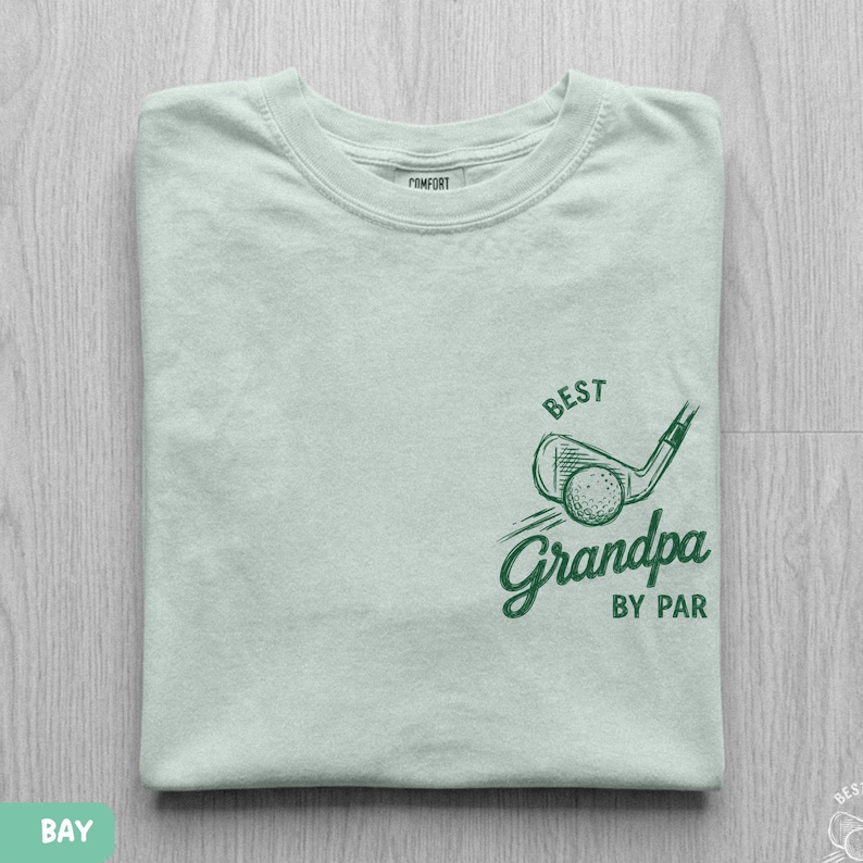 May include: A light green t-shirt featuring a dark green graphic of a golf club and ball with the text "BEST Grandpa BY PAR" printed on the front. The shirt has a crew neckline and a label on the inside collar that reads "COMFORT".