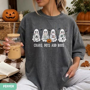 May include: A charcoal gray t-shirt featuring a graphic of four white ghosts holding mahjong tiles, with a small orange jack-o'-lantern in the center. Below the ghosts, the text reads "CRAKS, DOTS AND BOOS" in a white, clean, sans-serif font.