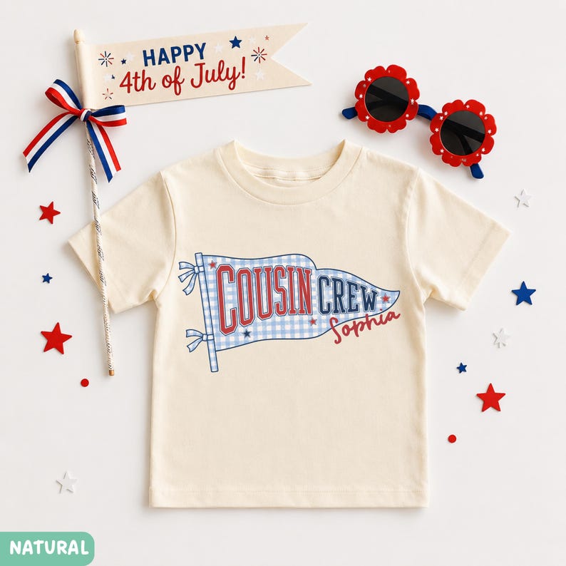 May include: A cream-coloured t-shirt with a graphic that reads "COUSIN CREW" in red and blue, with a gingham pattern. A wooden sign that says "Happy 4th of July!" is also in the image, along with red and blue star decorations and flower-shaped sunglasses.