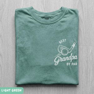 May include: A light green short-sleeve t-shirt featuring a white graphic on the chest. The design includes a golf club and ball illustration with the text "BEST" above and "Grandpa BY PAR" below in a stylized, hand-drawn white font.