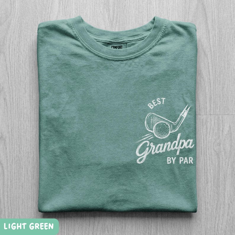 May include: A light green short-sleeve t-shirt featuring a white graphic on the chest. The design includes a golf club and ball illustration with the text "BEST" above and "Grandpa BY PAR" below in a stylized, hand-drawn white font.