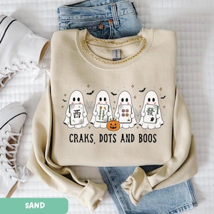 May include: A sand-colored crewneck sweatshirt featuring a graphic of four ghosts holding mahjong tiles, a small orange pumpkin, and spiderwebs. The text below the ghosts reads "CRAKS, DOTS AND BOOS" in black capital letters. The sweatshirt is layered over blue denim jeans.