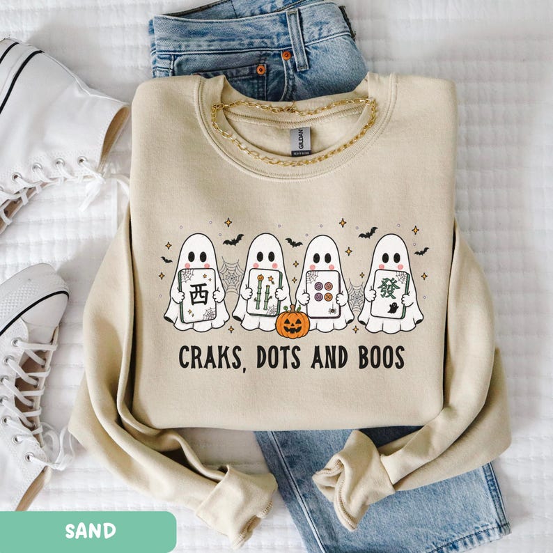 May include: A sand-colored crewneck sweatshirt featuring a graphic of four ghosts holding mahjong tiles, a small orange pumpkin, and spiderwebs. The text below the ghosts reads "CRAKS, DOTS AND BOOS" in black capital letters. The sweatshirt is layered over blue denim jeans.