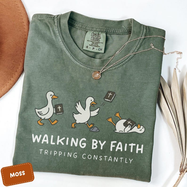 May include: Moss green t-shirt with a graphic of three cartoon ducks with Bibles. The text "WALKING BY FAITH TRIPPING CONSTANTLY" is printed below the ducks. A gold necklace with a pendant is draped on the shirt.