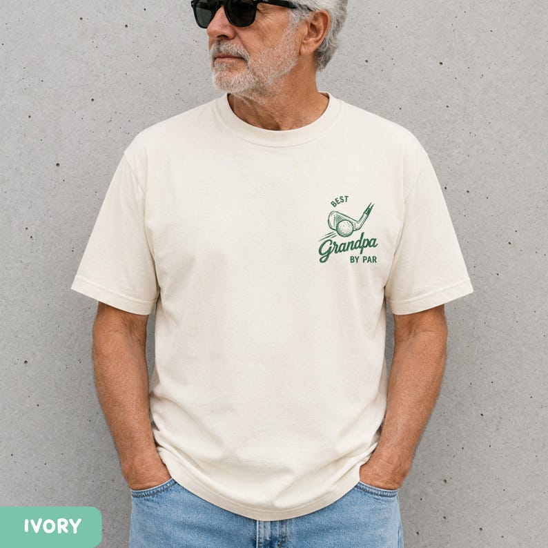 May include: An ivory short-sleeved t-shirt featuring a green graphic on the left chest. The design includes a golf club and ball illustration with the text "BEST" above and "Grandpa BY PAR" below in a stylized green script font.