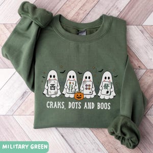May include: A military green crewneck sweatshirt featuring a graphic of four white ghosts holding mahjong tiles. A small orange jack-o'-lantern sits in front of the ghosts. The text "CRAKS, DOTS AND BOOS" is printed in white below the graphic.