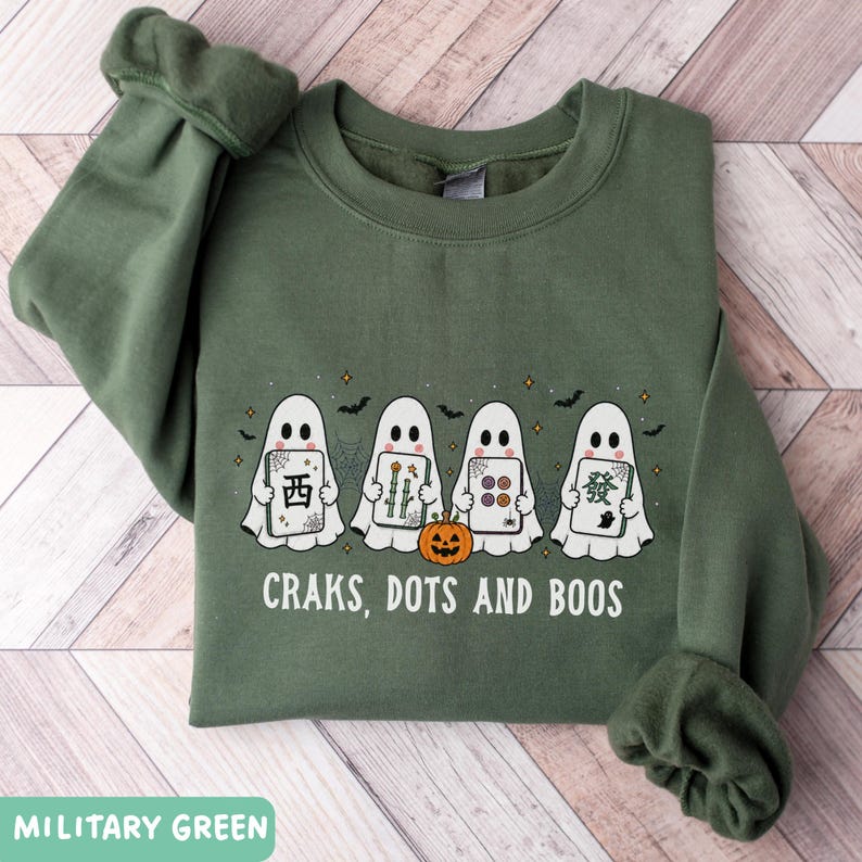 May include: A military green crewneck sweatshirt featuring a graphic of four white ghosts holding mahjong tiles. A small orange jack-o'-lantern sits in front of the ghosts. The text "CRAKS, DOTS AND BOOS" is printed in white below the graphic.