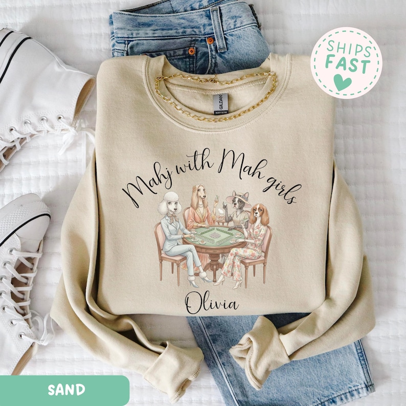 Op de afbeelding: Zandkleurige sweatshirt met de tekst "Mahj with Mah girls" en een illustratie van honden die mahjong spelen. De naam "Olivia" staat onder de illustratie. De sweatshirt wordt gecombineerd met een blauwe jeans en witte sneakers.
