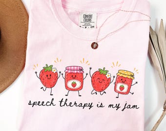 May include: A light pink t-shirt featuring a graphic of two smiling strawberries and two jars of strawberry jam with the text "speech therapy is my jam" printed in black. The shirt has a Comfort Colors brand label at the inner neckline.