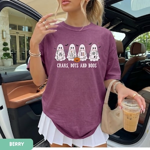 May include: A berry-colored t-shirt featuring a graphic of four white ghosts holding mahjong tiles and a small orange pumpkin. The text below the ghosts reads "CRAKS, DOTS AND BOOS". The shirt has a crew neckline and short sleeves.