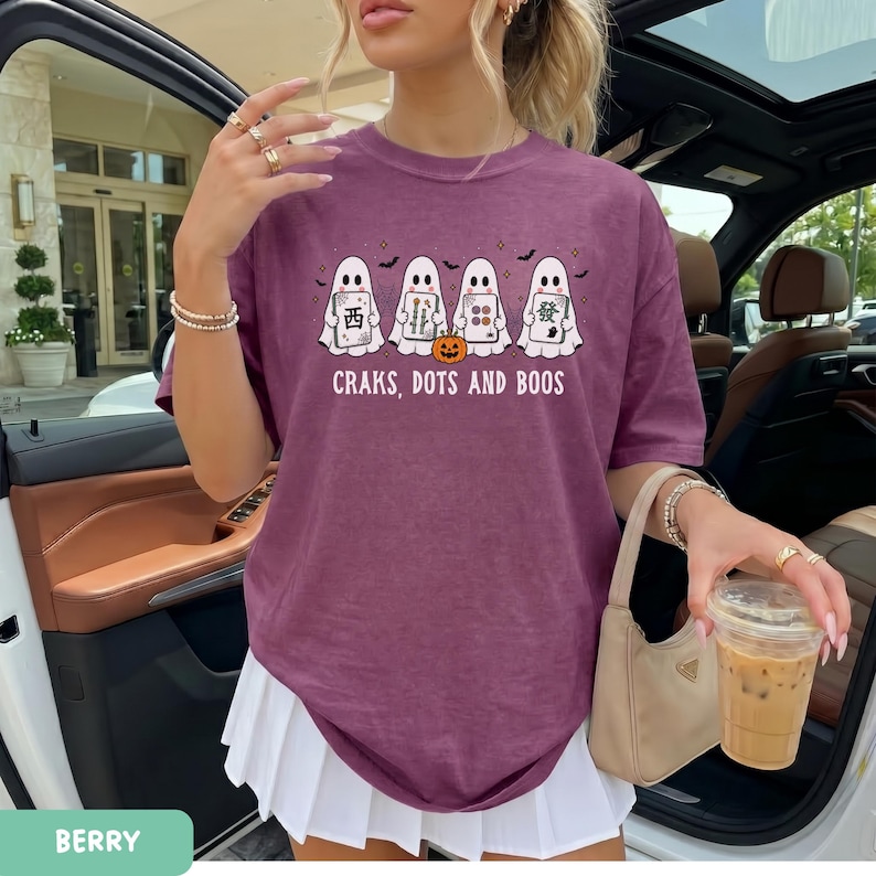 May include: A berry-colored t-shirt featuring a graphic of four white ghosts holding mahjong tiles and a small orange pumpkin. The text below the ghosts reads "CRAKS, DOTS AND BOOS". The shirt has a crew neckline and short sleeves.