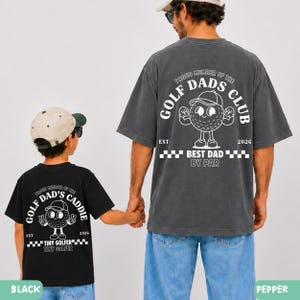 May include: Two matching t-shirts, one black and one pepper-colored, featuring a golf ball character with the text "Golf Dad's Club" and "Best Dad By Par." The black shirt says "Tiny Golfer". The shirts are worn by a father and child.