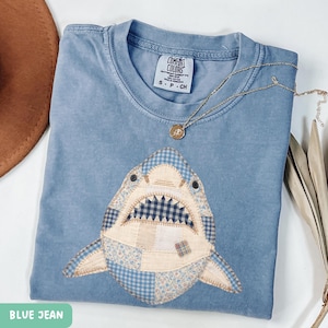 May include: A blue jean Comfort Colors t-shirt featuring a shark design. The shark is made of patchwork fabric in various blue patterns. A gold necklace with a pendant rests on the shirt. The shirt has a small tag that says "S - P - CH".