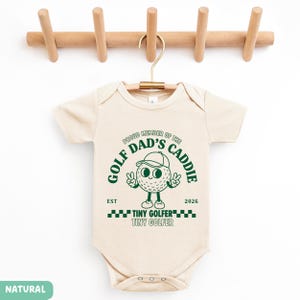 May include: A cream-colored baby onesie with the text "PROUD MEMBER OF THE DAD'S CADDIE" and a cartoon golf ball character. The onesie also has the words "TINY GOLFER" and the year "2026" printed on it. The onesie is hanging on a wooden hanger.