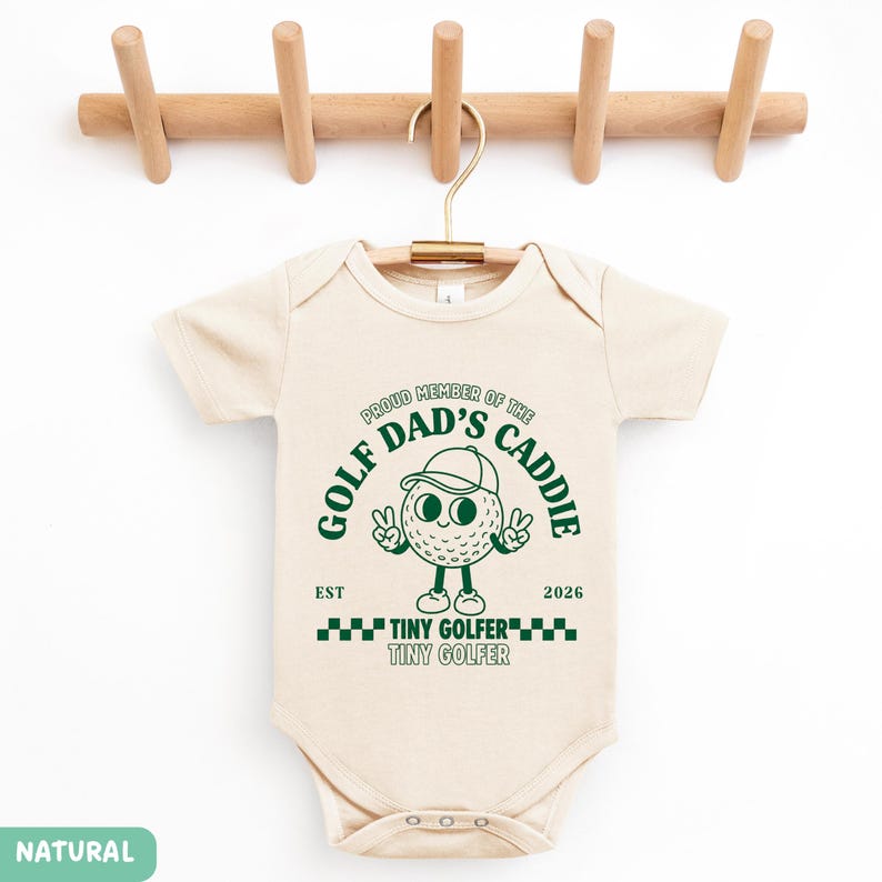 May include: A cream-colored baby onesie with the text "PROUD MEMBER OF THE DAD'S CADDIE" and a cartoon golf ball character. The onesie also has the words "TINY GOLFER" and the year "2026" printed on it. The onesie is hanging on a wooden hanger.