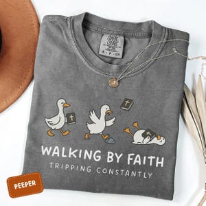 May include: A gray Comfort Colors t-shirt with a humorous graphic of cartoon ducks with Bibles. The text reads "WALKING BY FAITH, TRIPPING CONSTANTLY." A gold necklace and a brown hat are also visible.