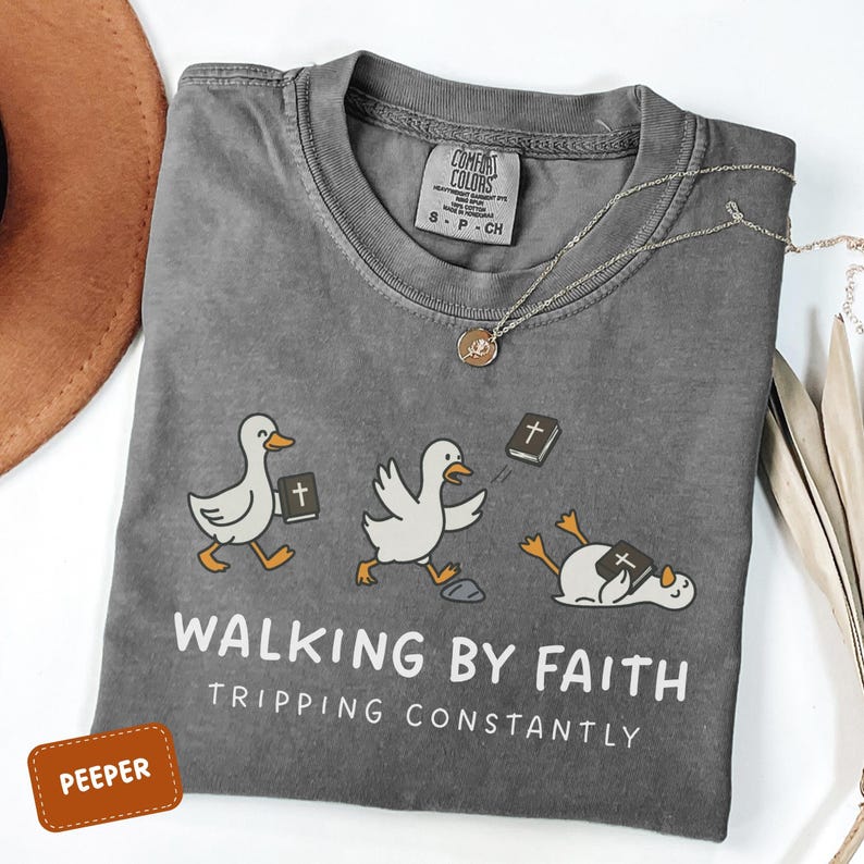 May include: A gray Comfort Colors t-shirt with a humorous graphic of cartoon ducks with Bibles. The text reads "WALKING BY FAITH, TRIPPING CONSTANTLY." A gold necklace and a brown hat are also visible.