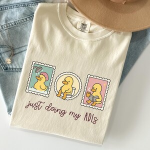 May include: Cream-colored t-shirt with a graphic of three duck stamps. Each stamp shows a yellow duck in different activities: showering, using the toilet, and reading with coffee. The text "just doing my ADLs" is below the stamps.