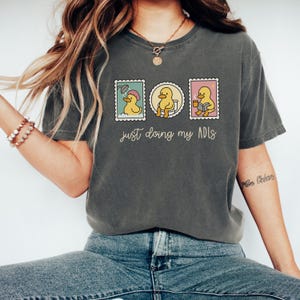 May include: Dark gray t-shirt with a graphic of three stamps featuring a yellow duck in various activities. The text "just doing my ADLs" is printed below the stamps. The shirt is made of a soft material.