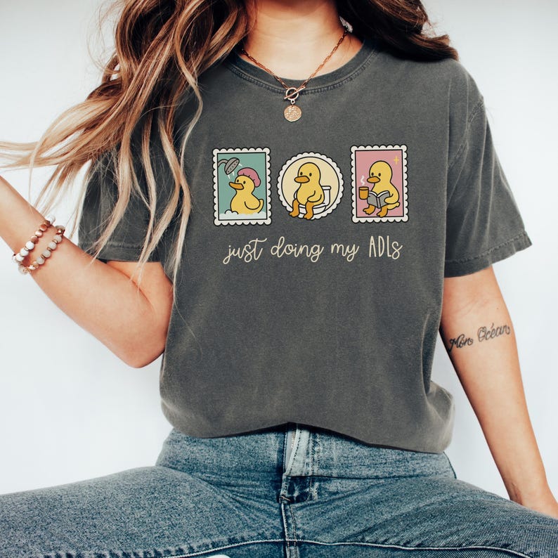 May include: Dark gray t-shirt with a graphic of three stamps featuring a yellow duck in various activities. The text "just doing my ADLs" is printed below the stamps. The shirt is made of a soft material.