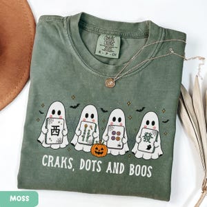May include: A moss green t-shirt featuring a graphic of four white ghosts holding mahjong tiles, a small orange pumpkin, and the text "CRAKS, DOTS AND BOOS" in white lettering. The shirt has a crew neckline and a Comfort Colors brand label.