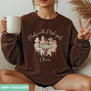 Op de afbeelding: Donker chocoladebruine sweatshirt met de tekst "Mahj with Mah girls" boven een illustratie van vier vrouwen die een spel spelen. De naam "Olivia" staat eronder. De sweatshirt heeft een ronde hals.