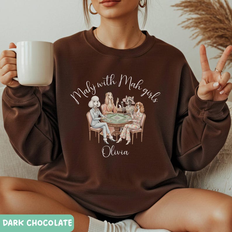 Op de afbeelding: Donker chocoladebruine sweatshirt met de tekst "Mahj with Mah girls" boven een illustratie van vier vrouwen die een spel spelen. De naam "Olivia" staat eronder. De sweatshirt heeft een ronde hals.