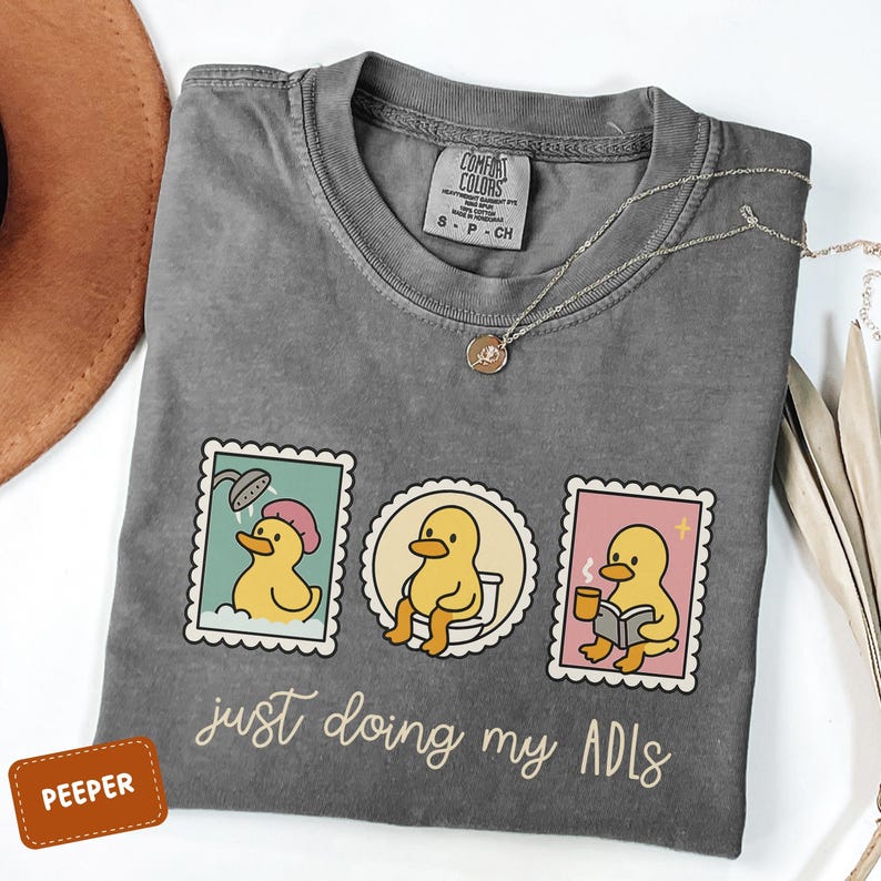 May include: A gray Comfort Colors t-shirt featuring three cartoon duck stamps. The stamps depict a duck showering, on the toilet, and reading. The text "just doing my ADLs" is below the stamps. A silver necklace and a brown hat are also visible.