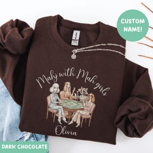 Op de afbeelding: Donkerchocoladekleurige sweatshirt met de tekst "Mahj with Mah girls" en een illustratie van honden die mahjong spelen. De naam "Olivia" staat onder de illustratie gedrukt. Een zilveren ketting ligt op de sweatshirt.