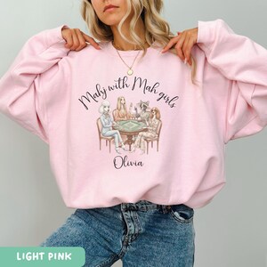 Op de afbeelding: Lichtroze sweatshirt met de tekst "Mahj with Mah girls" en de naam "Olivia". De afbeelding toont vier vrouwen die Mahjong spelen. De sweatshirt heeft een ronde hals.