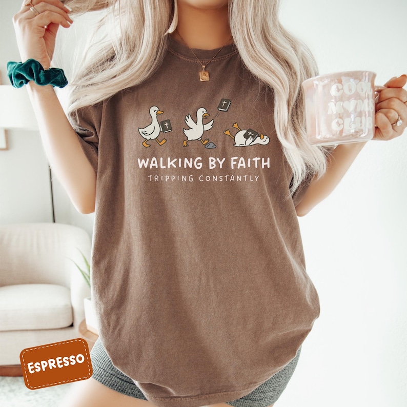 May include: Espresso-colored t-shirt with the text "WALKING BY FAITH TRIPPING CONSTANTLY" and a graphic of three ducks. The first two ducks are walking, and the third is lying down. The shirt is a comfortable, casual style.