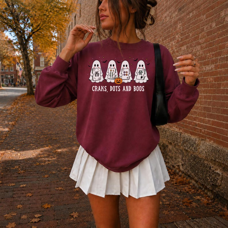 May include: A maroon long-sleeve crewneck sweatshirt featuring a graphic of four white ghosts holding mahjong tiles and a small orange jack-o'-lantern. The text "CRAKS, DOTS AND BOOS" is printed in white below the ghosts. The sweatshirt is paired with a white pleated mini skirt.