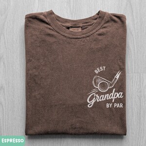 May include: A brown crew neck t-shirt featuring a white graphic on the lower right side. The graphic depicts a golf club and ball with the text "BEST" above and "Grandpa BY PAR" below the illustration. The shirt has a visible inner neck label.