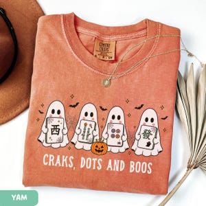 May include: A terracotta-colored t-shirt featuring a graphic of four white ghosts holding mahjong tiles, a small orange pumpkin, spiderwebs, and bats. The text "CRAKS, DOTS AND BOOS" is printed in white below the ghosts. The shirt has a crew neckline.