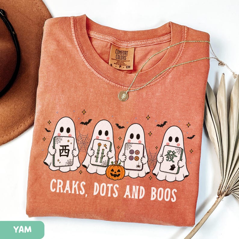 May include: A terracotta-colored t-shirt featuring a graphic of four white ghosts holding mahjong tiles, a small orange pumpkin, spiderwebs, and bats. The text "CRAKS, DOTS AND BOOS" is printed in white below the ghosts. The shirt has a crew neckline.