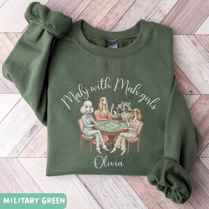 Op de afbeelding: Militairgroene sweatshirt met de tekst "Mahj with Mah girls" en een afbeelding van honden die mahjong spelen. De naam "Olivia" staat onder de afbeelding gedrukt. De sweatshirt is gemaakt van een zacht materiaal.