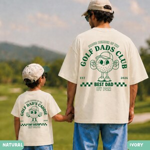 May include: Two ivory-colored t-shirts with green text and graphics. The shirts feature a golf ball character and the text "GOLF DAD'S CLUB" and "BEST DAD BY PAR". One shirt says "TINY GOLFER". The shirts are worn by a father and child.