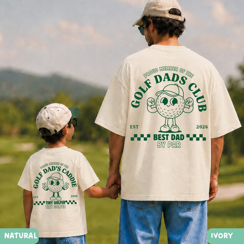 May include: Two ivory-colored t-shirts with green text and graphics. The shirts feature a golf ball character and the text "GOLF DAD'S CLUB" and "BEST DAD BY PAR". One shirt says "TINY GOLFER". The shirts are worn by a father and child.