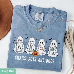 May include: A blue jean colored t-shirt featuring a graphic of four white ghosts holding mahjong tiles, a small orange pumpkin, spiderwebs, and bats. The text reads "CRAKS, DOTS AND BOOS" in white capital letters. A gold chain necklace rests on the collar.