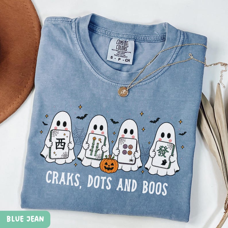 May include: A blue jean colored t-shirt featuring a graphic of four white ghosts holding mahjong tiles, a small orange pumpkin, spiderwebs, and bats. The text reads "CRAKS, DOTS AND BOOS" in white capital letters. A gold chain necklace rests on the collar.
