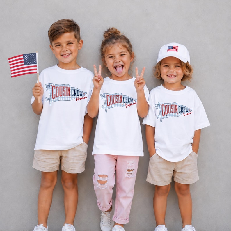 May include: Three children wearing white t-shirts with a "COUSIN CREW" flag design and individual names. One child holds an American flag, and another wears a baseball cap with the American flag. The children are wearing khaki shorts and pink trousers.