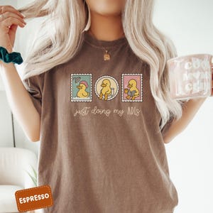 May include: Espresso-colored t-shirt featuring a graphic of three duck stamps. Each stamp depicts a duck in different activities: showering, using the toilet, and reading. The text "just doing my ADLs" is below the stamps.
