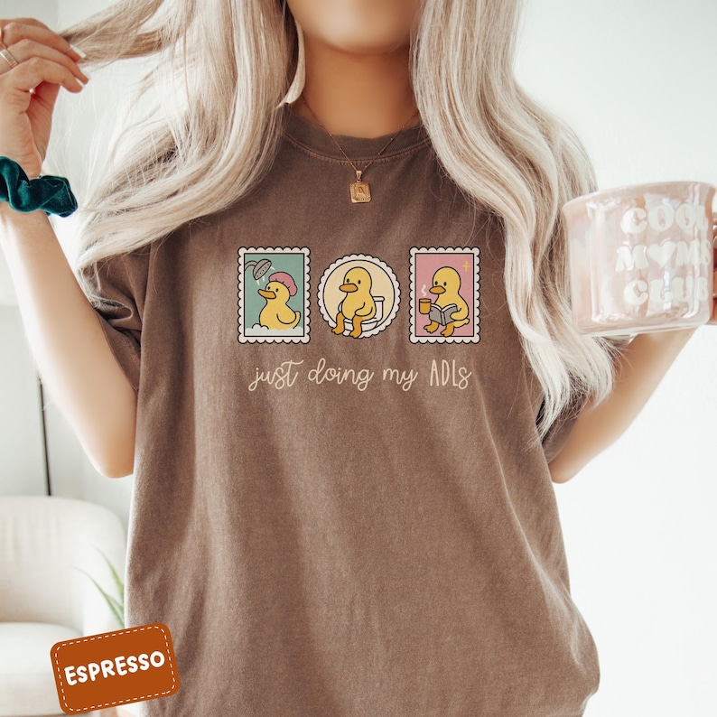 May include: Espresso-colored t-shirt featuring a graphic of three duck stamps. Each stamp depicts a duck in different activities: showering, using the toilet, and reading. The text "just doing my ADLs" is below the stamps.