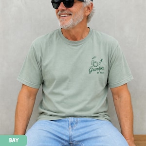 May include: A sage green short-sleeve t-shirt featuring a dark green graphic on the left chest. The graphic depicts a golf club and ball with the text "BEST" above and "Grandpa BY PAR" below in a stylized script and block font combination.