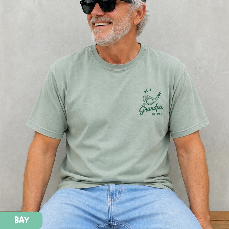 May include: A sage green short-sleeve t-shirt featuring a dark green graphic on the left chest. The graphic depicts a golf club and ball with the text "BEST" above and "Grandpa BY PAR" below in a stylized script and block font combination.