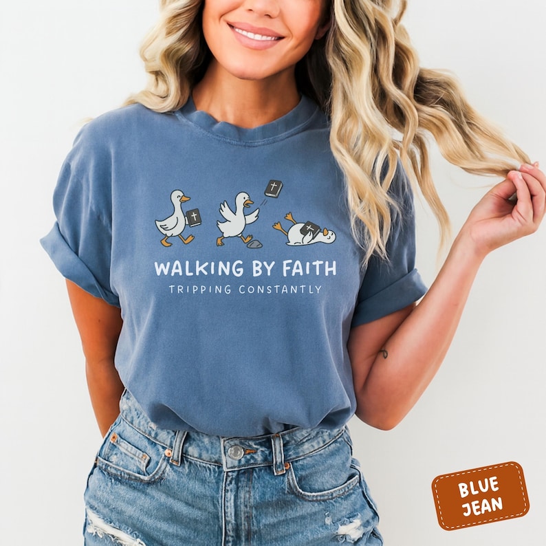 May include: Blue jean-colored t-shirt with the text "WALKING BY FAITH TRIPPING CONSTANTLY" and a graphic of three ducks with Bibles. The first two ducks are walking, and the third is lying down. The shirt is a relaxed fit.
