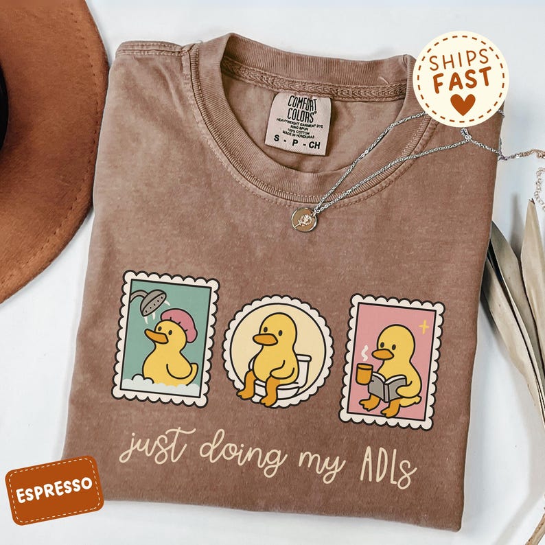 May include: Espresso-colored t-shirt with a graphic of a yellow duck in various daily activities, including showering, using the toilet, and reading. The text "just doing my ADLs" is below the graphic. A silver necklace is draped on the shirt.