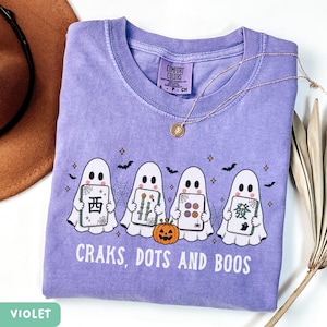 May include: A violet t-shirt featuring a graphic of four white ghosts holding mahjong tiles, a small orange jack-o'-lantern, spiderwebs, bats, and stars. The text "CRAKS, DOTS AND BOOS" is printed in white below the ghosts. The shirt has a Comfort Colors label.