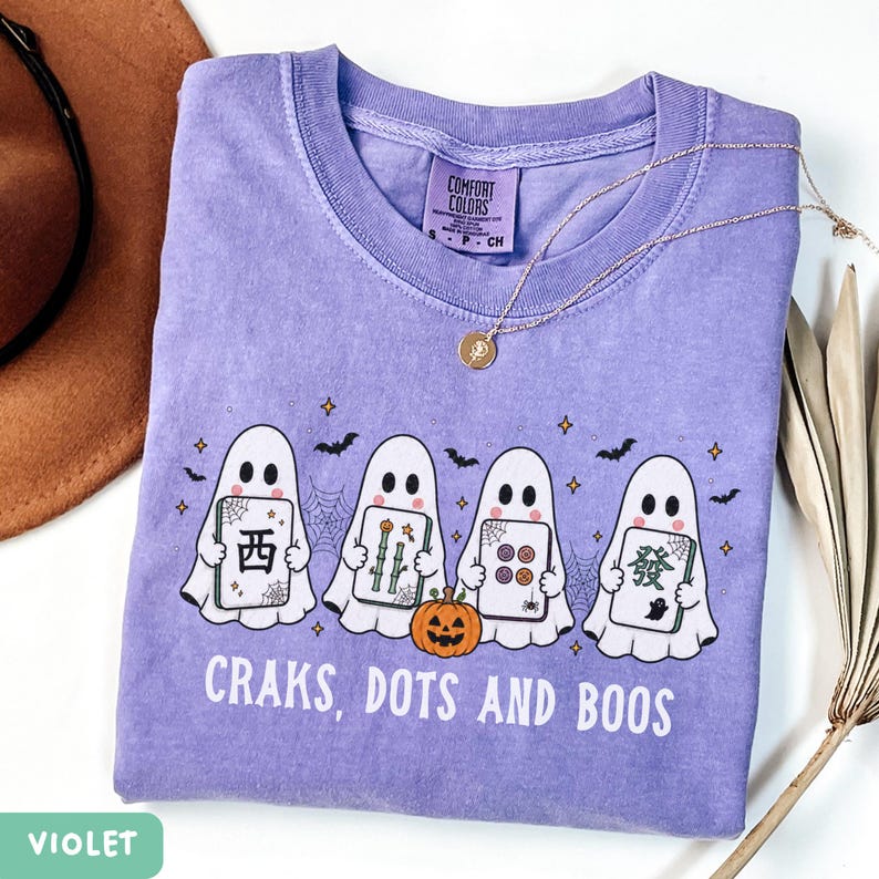 May include: A violet t-shirt featuring a graphic of four white ghosts holding mahjong tiles, a small orange jack-o'-lantern, spiderwebs, bats, and stars. The text "CRAKS, DOTS AND BOOS" is printed in white below the ghosts. The shirt has a Comfort Colors label.
